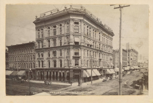 A view of the Tabor Block, designed by Frank E. Edbrooke, at 16th (Sixteenth) and Larimer Street in Denver, Colorado. Horses and carriages are parked in the street. Signs on the Tabor building read "Designated Depository of the United States", "Bank", "Rose & Co. Photographers", "Gurlly Bros." and "Winne & Co.". A sign on the next building reads "Hotel Brunswick".