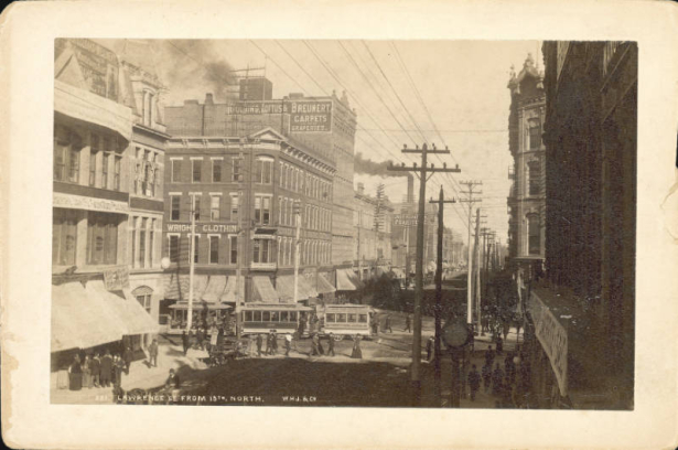 A view of Lawrence and 15th (Fifteenth) streets in Denver, Colorado; shows pedestrians, cable cars, utility poles, smokestacks, and commercial buildings. Signs read "Wright Clothing" and "Golding, Loftus & Breunert Carpets and Draperies.
