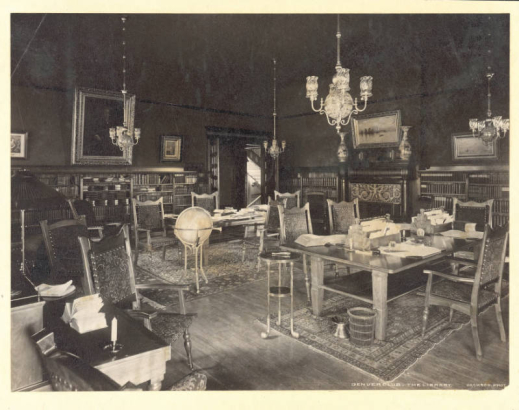 A view of the library in the Denver, Club at 17th (Seventeenth) and Glenarm in Denver, Colorado. The interior has upholstered wooden chairs, tables, bookcases, paintings in gilded frames, chandeliers, and a fireplace.