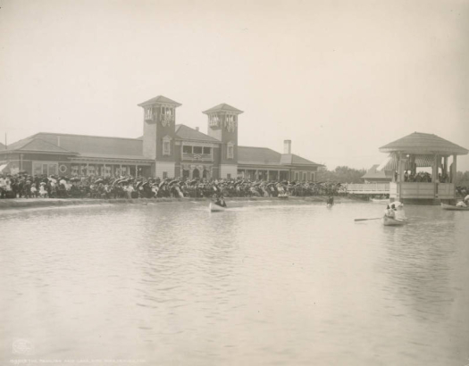 View of the pavilion near City Park (Ferril) Lake in City Park, Denver, Colorado. The Spanish Colonial Revival style building has towers with tile roofs and an arcade. People sit on chairs at the shore of the lake with parasols as groups in rowboats go by.  A band can be seen in the bandstand on the lake.