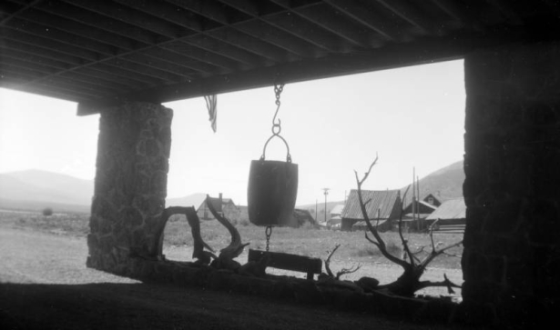 View from the Ore Bucket Lodge in Crested Butte, Colorado; shows the silhouettes of an ore bucket hanging from a rafter on a chain, and a large tree branch laying under the bucket; two large stone columns and a roof form a frame for the two objects; image taken from shadow into light; residences and mountains in the background.