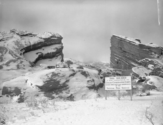 View of a sign near the unfinished Red Rocks Amphitheater in Red Rocks Park, a Denver Mountain Park, in Jefferson County, Colorado. The sign reads: "This Theater is Under Construction by the Civilian Conservation Corps Under Direction and Supervision of the National Park Service." Shows snow covered rock formations, concrete seats, and stone buildings.