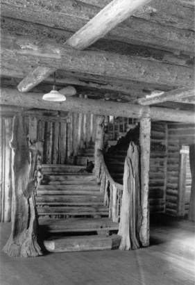 View of a rustic wooden staircase in Enos Mills' Long's Peak Inn in Rocky Mountain National Park in Colorado. The staircase has weathered logs as newel posts, and knotted pine balusters. The building has log walls and a wood floor. Some of the walls are chinked. The ceiling is constructed of bark covered pine log beams. An electric light hangs over the staircase.