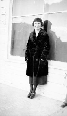 Grace Bedford poses in Keota, Weld County, Colorado, wearing  black shoes, stockings, skirt, and fur coat. She has black hair, cut in a "bob." Windows in a frame wall are behind her.
