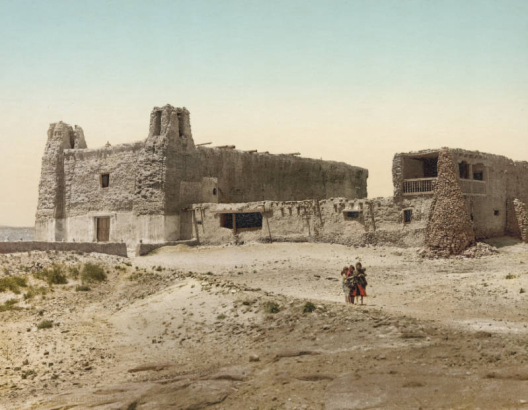 View of San Esteban del Rey de Acoma, a mission church, and the convento (convent) at Acoma Pueblo, New Mexico. The buildings are constructed of adobe and stone. The church has two bell towers; the two-story convent is attached to the mission building and has a balcony with a wooden railing and stone buttresses. Girls stand near the buildings.