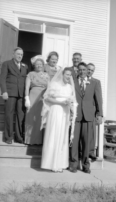 Posing on the church steps in Keota, Weld County, Colorado, This wedding party wears suits, ties, gloves, boutonnieres, corsages, lace hats and veils. The groom has several teeth missing.