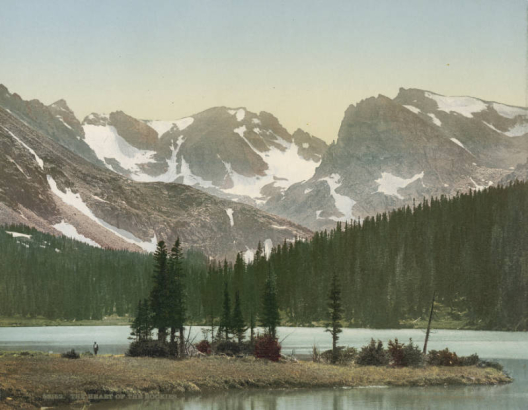 View of Long Lake and Navajo Peak, Apache Peak, and Shoshoni Peak, in the Indians Peaks in Boulder County, Colorado. A man stands on a spit with trees on the lake.