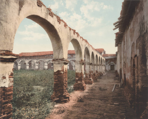 View of an arcade at Mission San Juan Capistrano in San Juan Capistrano, California. Plants grow from the top of a colonnade, and rubble is piled against a wall.  Plants grow wild in the central patio of the mission. The adobe walls and flagstone arches of the mission have missing plaster. The buildings have red clay tile roofs.