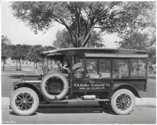 View of a Ford Model T delivery truck in City Park, Denver, Colorado. Lettering on the truck reads: "The Colorado House" and "The W.H. Kistler Stationery Co. 1636-46 Champa St." Two men sit in the truck. The bed of the truck has wire mesh and rolled-up canvas covers. Packages are in the back of the truck. A metal cylinder is mounted on the running board of the truck. Trees, sidewalks and people are in the distance.