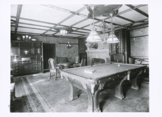 View the billiard room at Cleveholm (Osgood Castle), the home of John Cleveland Osgood, located in Redstone (Pitkin County), Colorado. The room has a large, carved billiard table, a fireplace flanked by two glass fronted cases, a carved sideboard and a table and chairs. A bronze chandelier with two glass shades hangs over the billiard table. An oriental rug is on the floor.