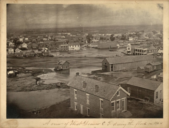 View of flooded Cherry Creek and inundated buildings in Denver, Colorado, Shows brick and frame houses and commercial buildings. Signs read "American House," "Commonwealth," and "J.G. Vawter & Co." The South Platte River and the Front Range are in the distance.
