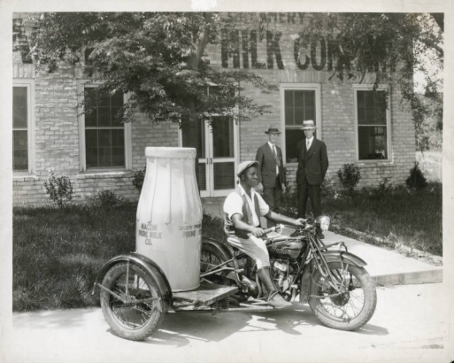 An African American man rides an Indian Motorcycle with a Winter-Weiss Company platform sidecar in front of the Macon Pure Milk Company in Macon, Georgia. He wears a cap, vest, gaiters add boots. An advertising model of an oversized milk bottle is on the sidecar's platform. Lettering on the bottle reads: "Macon Pure Milk Co." Two men in suits and hats look on.