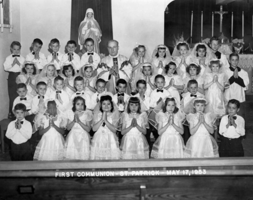 Holding prayer books with hands poised for prayer, This communion class (one Black student) in Holyoke, Phillips County, Colorado poses in white shirts and taffeta dresses. In vestments, a priest is in the  back row; behind them are an altar with candles and flowers, and a likeness of the Virgin Mary.