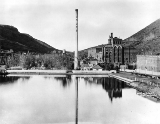 View east of the Adolph Coors Company Brewery, between Table Mountain, Golden, Jefferson County, shows lake in front of the multi-story buildings, tall "Coors" smokestack and wooden fence.