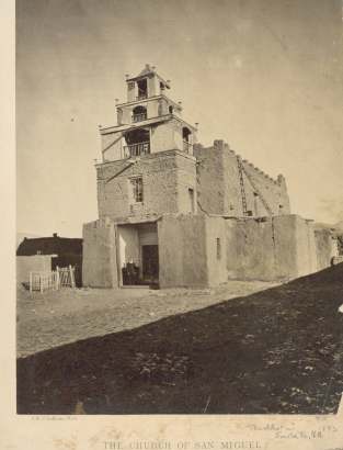 Men pose in the entrance or narthex of the adobe Church of San Miguel in Santa Fe (Santa Fe County), New Mexico. One man, a priest, wears a cassock. The church has tapered towers with wooden railings and battlements along the roof line of the nave. Shows wooden lintels above the entrance and a wooden ladder at the side of the church.