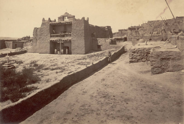 View of an adobe Nuestra Señora de Guadalupe mission church at Zuni Pueblo, New Mexico. Native American (Zuni) men pose in the entrance and on a second floor balcony. The church has two bells in a central belfry, worn battlements on towers, and vigas near the roof line of the nave. A cross and altar are in the walled courtyard. A man, probably a member of the survey crew, leans against an adobe wall.