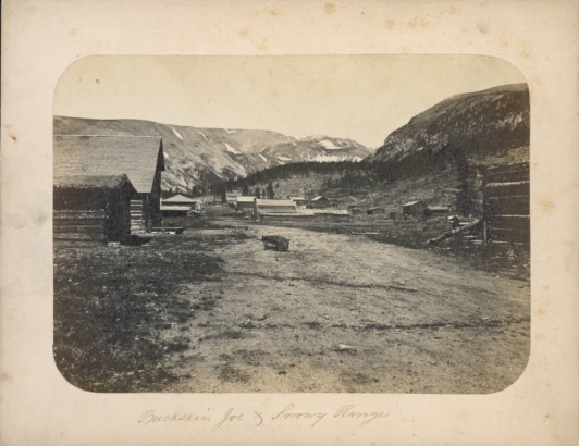 View of the dirt main street in Buckskin Joe (Park County), Colorado. Shows log and wood frame buildings that include cabins, sheds and commercial structures. One cabin has a sod roof. A wheelbarrow is in the middle of the dirt road. Probably the Mosquito range is in the distance.