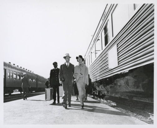 A man and woman passenger prepare to board the Southern Pacific Coast Daylight at the 3rd & Townsend Street Station in San Francisco, California. An African American baggage porter walks behind them with two suitcases. Shows the aluminum exterior of the streamliner.