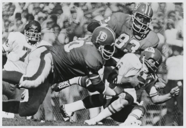 Two Denver Broncos professional football players tackle an opponent. Spectators are in the background.