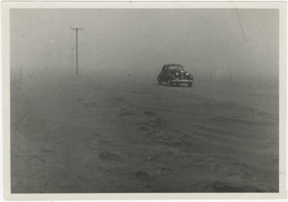 A two-door sedan approaches amid blowing dust. Telephone poles are to the sides.