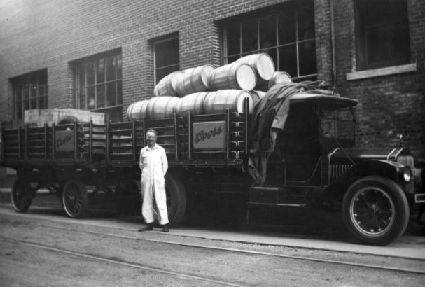 Len Vogel stands in his white overalls in front of his Coors delivery truck at the Adolph Coors Brewery, Golden, Jefferson County, Colorado. The truck has an open frame bed loaded with wooden barrels and another trailer attached with wooden crates. A canvas covering for the load  is on top the truck cab.
