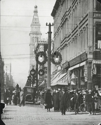 Pedestrians walk along bustling Sixteenth Street around noon at the intersection of Stout Street in the City and County of Denver, Colorado.  Christmas wreaths hang from the lightposts.  Cars and bicycles are parked along the street.  Landmarks include the Daniels and Fisher Tower.