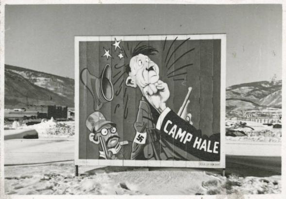 Billboard in the snow outside of Camp Hale, a U.S. Army training camp for World War II mountain soldiers, near Tennessee Pass, Colorado. Image on the billboard is of a clenched fist with "Camp Hale" printed on the sleeve punching a caricature of Adolph Hitler (wearing a military uniform with a swastika on the sleeve and holding a rifle) in the jaw.  A caricature of a Japanese soldier next to Hitler wears a military uniform and holds a rifle.