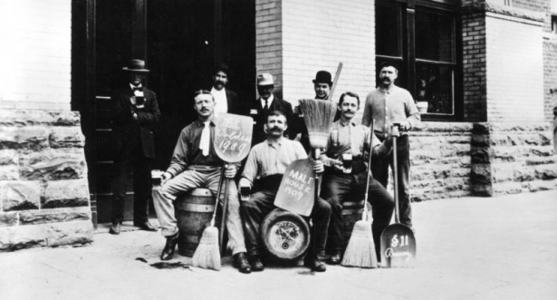 Group of men pose outside the Adolph Coors Company brewery, Golden, Colorado; they hold long handled whisk brooms, mugs of beer, and shovels with "Wet Golden 1909," "Malt House 1909" and "Section 11 Brewery," lettered on them. Lennard Vogel sits on a wooden barrel keg on the left, Alphonse Thuet stands in the back right.