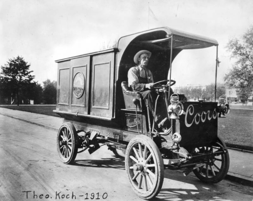 Theo Koch drives an Adolph Coors Golden Brewery delivery truck, in Golden, Jefferson County. The wood panel Frayer - Miller truck has the steering wheel on the right side, narrow tires and lantern headlights.