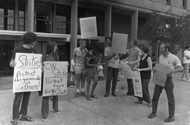 Students at the University of Colorado at Boulder carry hand lettered signs: "Strike! protest the genocide in Vietnam," "On strike protest Nixon and his war in insanity," "On Strike no more war," "Stop the atrocities in Asia and on campus on strike," "On strike for peace," "End the war shut it down," "Shut it down for your own sake." Campus buildings are in the background.