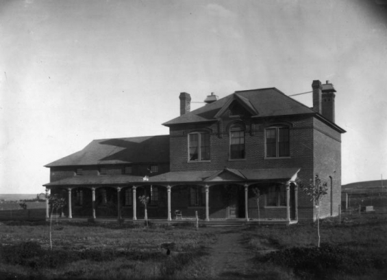 Native grass and new trees surround the Hospital (built in 1884) at the University of Colorado at Boulder. The two-story brick structure has an open porch, wood trim, a truncated cottage roof, and braced chimneys. A dormer tops the central arched window, and the east wing has a lower roof and small second-story windows. Prairie fills the background.