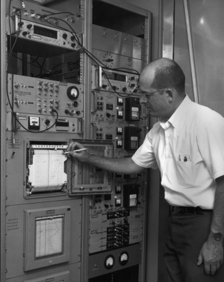 At the National Bureau of Standards in Boulder, Colorado, John Milton targets a rolling graph paper with a mechanical pencil. The date on the paper is: "Jul 23 1968." The glass, metal, and rubber gasket cover is opened, and the surrounding metal frames have electronic components mounted in them. Gauges, toggle switches, dials, digital (mechanical) readouts, and RF cables front each of the instruments.