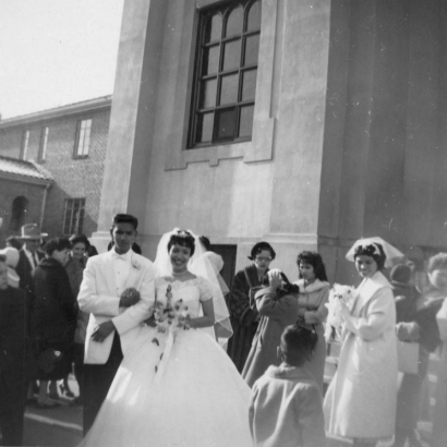 Juanita Ramos née Solano, poses with her husband, Tony Ramos and members of their wedding party in front of St. Cajetan's Catholic Church, 1190 Ninth (9th) Street in the Auraria neighborhood, Denver, Colorado. The bride wears a wedding dress with short sleeves and a full length skirt, she holds a bouquet. The groom holds her arm and wears a white coat, bow tie and dark pants. A woman in a coat, holds a bouquet and wears a veil on her head.