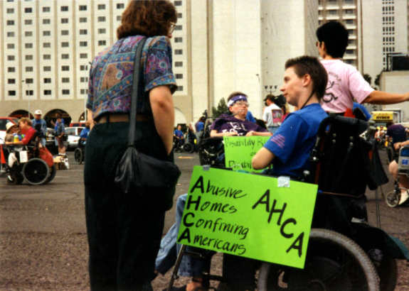 Robin Stephens protesting with ADAPT against the American Health Care Association's 1994 Las Vegas convention. Stephens has a handwritten sign taped to her wheelchair that reads "Abusive Homes Confining Americans AHCA." Laura Hershey is visible in the background. Photographer unknown.