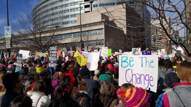 Large group of protesters by the Wellington Webb Building. Sign in the foreground "Be the Change"