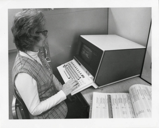 Photograph of an unidentified administrative assistant for the Denver Public Schools offices.  In the photograph she is sitting at an early computer entering in numbers for budget purposes.