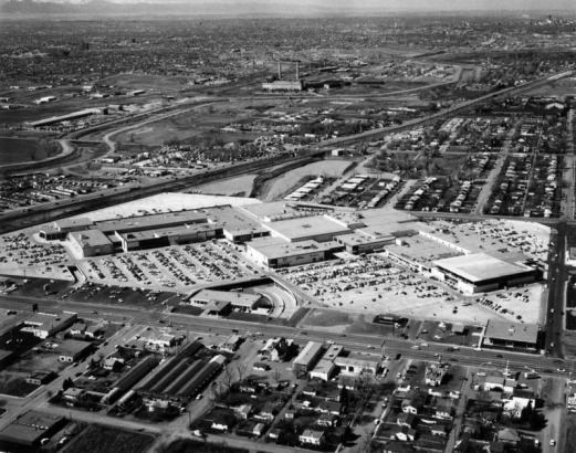 The zig-zag floor plan of Cinderella City shopping center, opened in 1968, is visible in an aerial view. The huge complex of connected two- story buildings and parking in Englewood, Colorado, is between Santa Fe Drive and Elati Street, and Hampden Avenue and Floyd Ave.; residential neighborhoods surround the area. Signs on buildings include: Cinema," "Woolworth's," "Western Federal Savings," "Englewood."