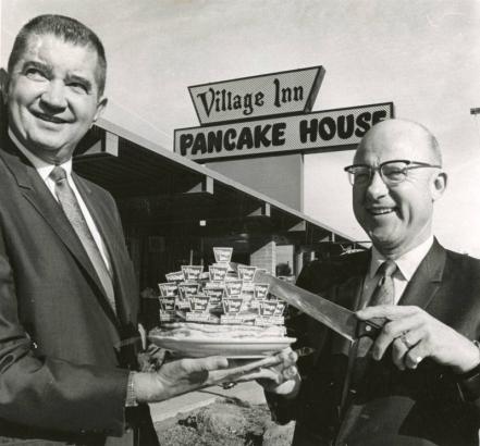 Merton S. (Andy) Anderson, left, vice-president, and Jim P. Mola, president of the Village Inn pancake house restaurants, celebrate the 10th birthday of their franchise operation with a birthday cake made of pancakes. The restaurant behind them at 8855 E. Colfax Ave., was the first in the group which now numbers 37 stores.