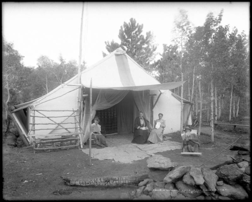 View of a campsite in the Rocky Mountains of Colorado; large canvas tent is supported by wooden posts with awning extended over front door to make a "covered porch;" three women sit underneath awning (one weaves reed or cane, one reads, one needlepoints); a man sits at edge of "porch" reading McClure's Magazine dated September, 1906; shows blankets on  ground for carpet, mosquito netting over tent doorway, and split rail or log bench; a burned log and rocks form firepit ring, center foreground.