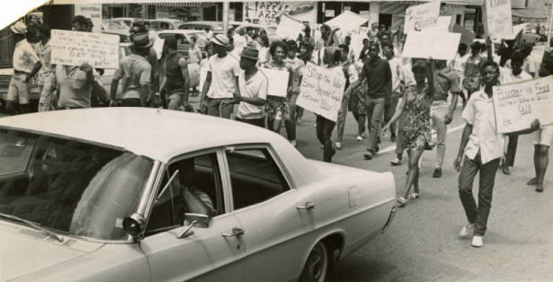 From back of photograph: Demonstrators march against traffic during trek Saturday to Police Bldg. Lt. Jerry R. Caroccia drives police car.