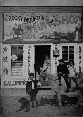 Children pose with a dog and a burro in blinders on a dirt street in Boulder, Colorado. A woman in a plaid blouse stands in the door of the storefront behind them. The sign above depicts a country scene with a waterfall and a troubadour on horseback blowing a horn; ornate letters read: "Rocky Mountain Paint Shop." Lettering below is in Chinese, German, and French, saying "Paint Shop." A birdcage hangs next to the entry; paintboxes and art comprise the window display.