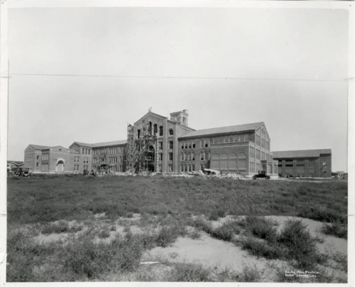 A black and white photograph of the exterior of South High School during construction in 1925.  The photograph is taken from across a field.  There is scaffolding in front of the future entrance to the school.  There are various cars visible around the building. Taken at 1700 East Louisiana Avenue, Denver, Colorado, 80210.