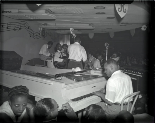 Interior photograph featuring Lionel Hampton playing the piano for a show at the Rainbow Ballroom at 38 East Fifth Avenue (E. 5th Ave.), Denver, Colorado. The rest of the band is in front of the piano, including the guitar and bass players.