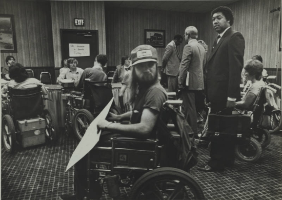 McDonald's executives Dennis Morris [with briefcase in hand], Don Fowler [with books], and McDonald's attorney [standing at back] wait for a meeting with people who use wheelchairs after a protest at the McDonald's on Colfax Avenue and Pennsylvania Street on May 11, 1984.