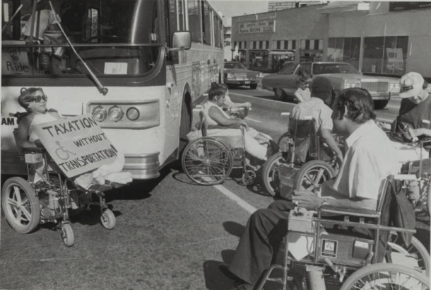 A group of protesters who use wheelchairs stop traffic to raise awareness of the lack of accessibility regarding the Regional Transportation District (RTD) busses. A sign on the left reads: "Taxation without transportation!" with the handicapped symbol drawn on the poster. 