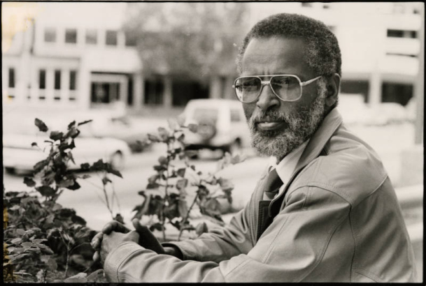 Tsegaye Hailu, who immigrated to the United States from Ethiopia, is now a hydrogeologist at J.F. Sato's engineering firm. He sits with his hands clasped facing the camera. He is wearing sunglasses.