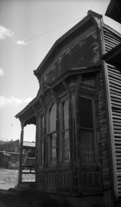 Detail view of a projecting bay window with sash windows in a house with gingerbread trim in Leadville, Colorado. The house has weathered  clapboard siding and a bay roof with decorative brackets continues over the front of the side porch. The center front window has a pointed arch, turned posts between windows, and a kickplate with three arches under each window sill. Lace curtains can be seen in the windows.