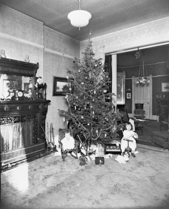 Interior view of a living room in a house in (probably) Denver, Colorado; shows small children and toys by a decorated Christmas tree. Shows: fireplace, toy piano, stuffed animals, and doll.