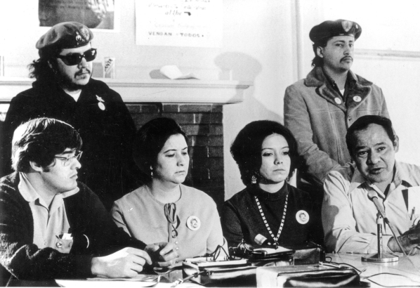 Leaders seated at the table include Joe Gonzales, Geraldine Gonzales, Nita Gonzales and John Haro speaking into a microphone. The two women wear buttons that have a photo of Chicano civil rights leader Rudolfo &quot;Corky&quot; Gonzales. Standing behind the group, possibly Brown Berets, are an unknown man with sunglasses and Eddie Montour of Pueblo, Colorado.