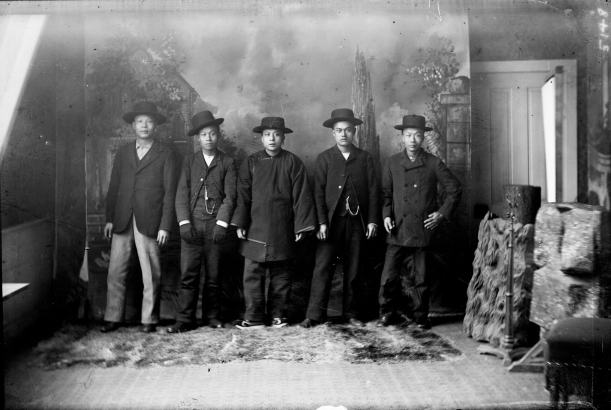 Studio portrait of Chinese-American men who include Wa Chin and Tang Ya-Shun in Georgetown (Clear Creek County), Colorado. Wa Chin wears a long shirt and wide-brimmed hat. Tang Ya-Shun wears a suit and watch chain with a charm.